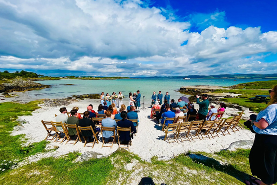 View of guests sitting on sandy beach for seaside ceremony